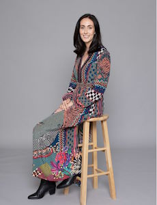 Portrait Photograph of Nikki Erlick. Grey background with a female looking towards the camera while sitting on a stool